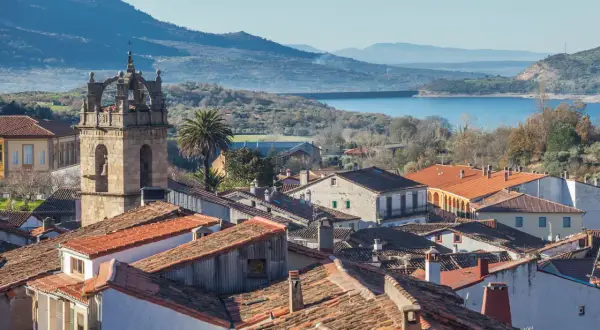 Vista de tejados y un campanario en las Hurdes, con un embalse y montañas al fondo.