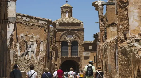 Gente camina por las ruinas de Belchite Viejo, un pueblo fantasma con edificios derruidos y una iglesia en el centro.