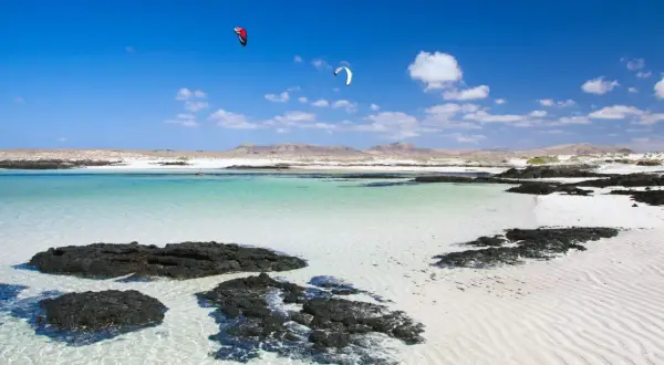 Aguas turquesas y arena blanca en Fuerteventura, con kitesurfistas en el horizonte. Un paraíso entre los 10 sitios para visitar en Fuerteventura.