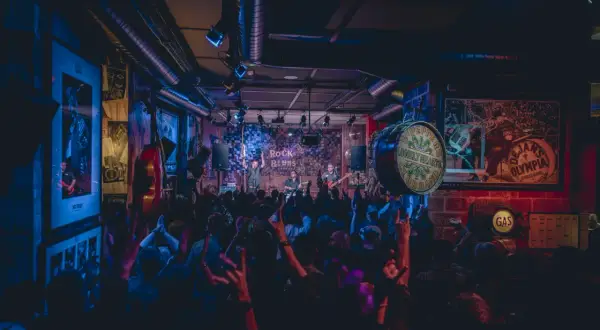 Una banda toca rock y blues en un local con luces de neón. El público levanta las manos, creando una atmósfera vibrante, como si estuvieran en una gran casa en Zaragoza.