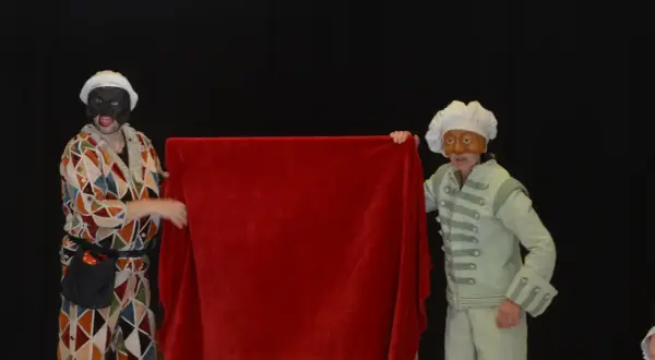 Dos actores con máscaras y trajes de época, uno con un traje de rombos y otro con un traje verde pálido, presentan una tela roja. Parecen parte de una obra de teatro de **els joglars zaragoza**.