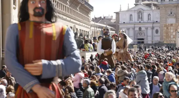 Gigantes y cabezudos desfilan entre una multitud en una plaza. Las figuras de cartón piedra, algunas con coronas, son el centro de atención.