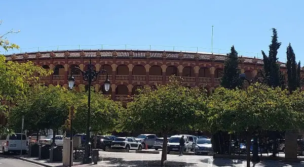 La plaza de toros de Zaragoza, con su arquitectura de arcos y balcones, se alza tras una hilera de árboles frondosos y un aparcamiento con varios coches.