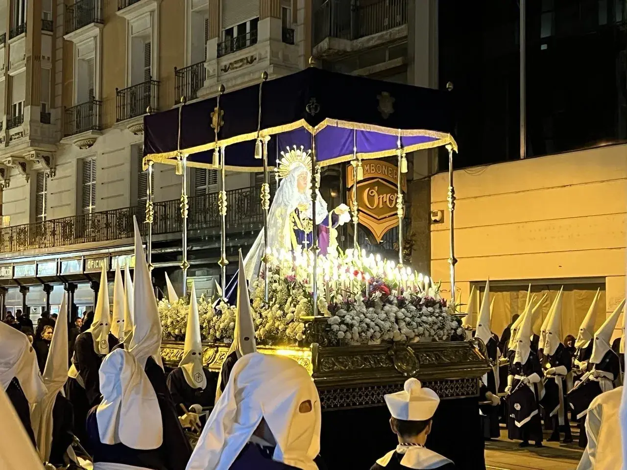 Procesión del Santo Entierro en Zaragoza. Figuras religiosas iluminadas por velas, rodeadas de nazarenos con túnicas y capirotes blancos.
