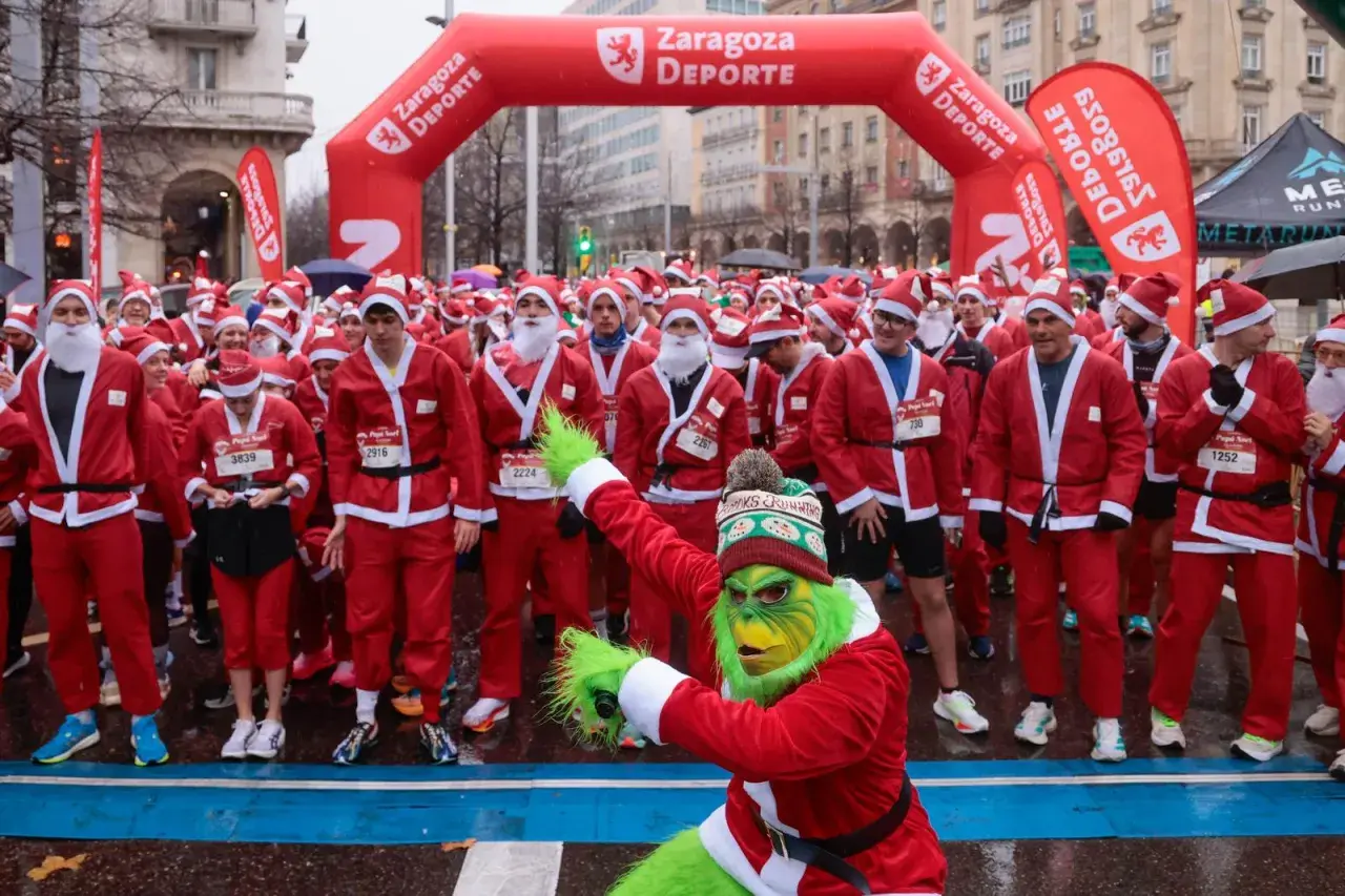 Multitud de personas disfrazadas de Papá Noel en Zaragoza, listas para una carrera. Un Grinch verde se une a la fiesta.