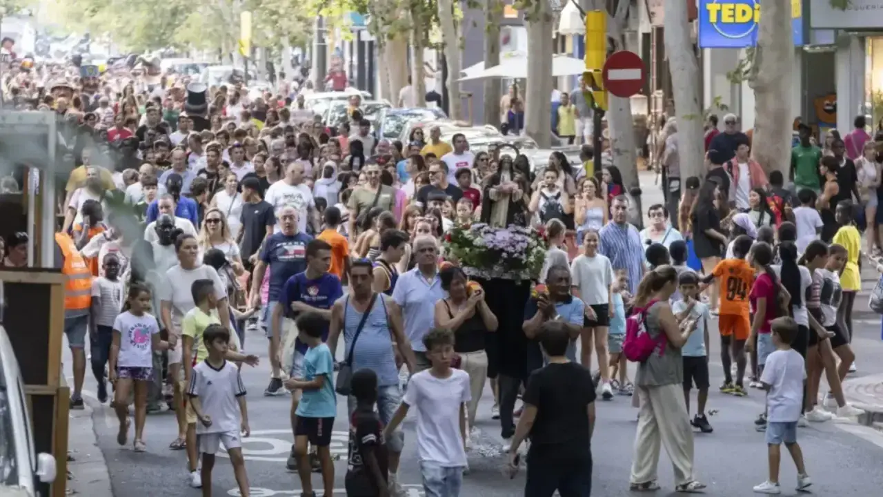 Multitud celebra las fiestas del barrio Las Fuentes en Zaragoza. Una procesión avanza por la calle con gente de todas las edades.