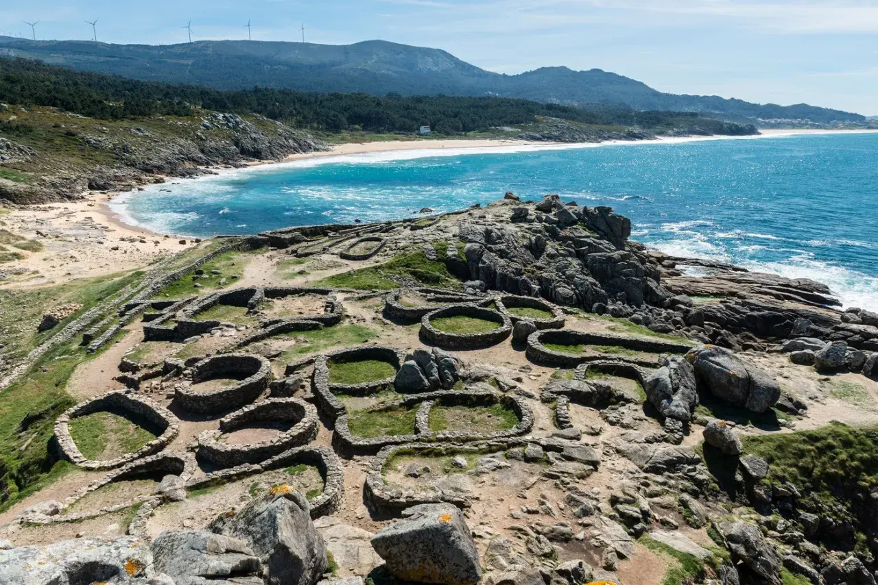 Ruinas circulares de un castro gallego, un lugar histórico que ver en A Coruña, con el mar azul y la costa de fondo.