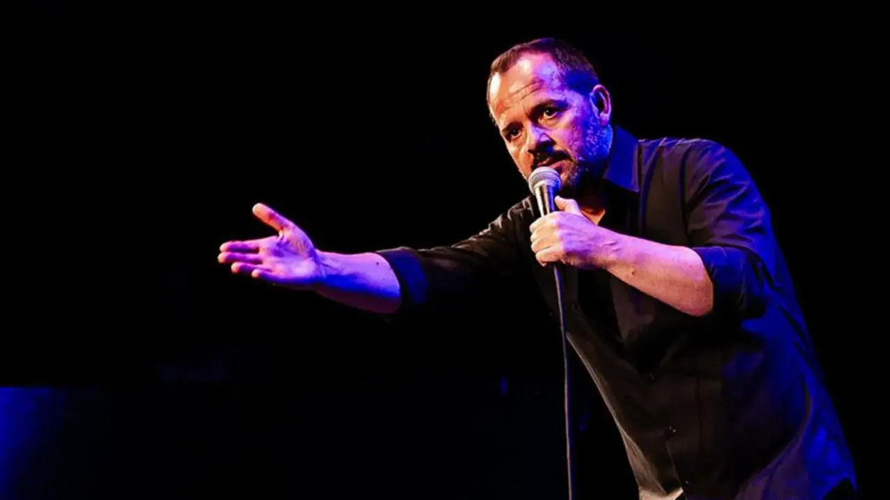 Ángel Martín Zaragoza, comediante, habla al micrófono con gesto expresivo y mano extendida, iluminado por luces azules y púrpuras.