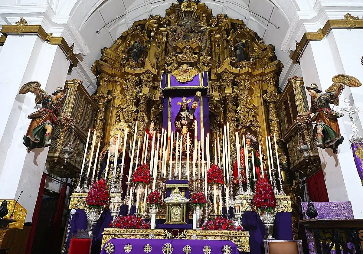 Un altar barroco con imágenes religiosas, velas y flores rojas. Destaca una figura central de Cristo, que parece ser un quinario, rodeado de figuras de la Virgen y otros santos.
