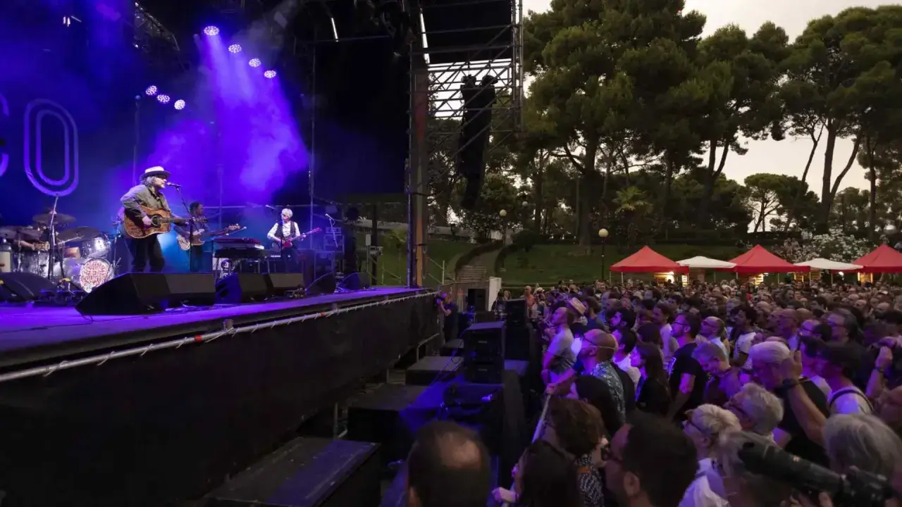 Concierto en un jardín de invierno con pilares de escenario y una multitud disfrutando bajo luces púrpuras.