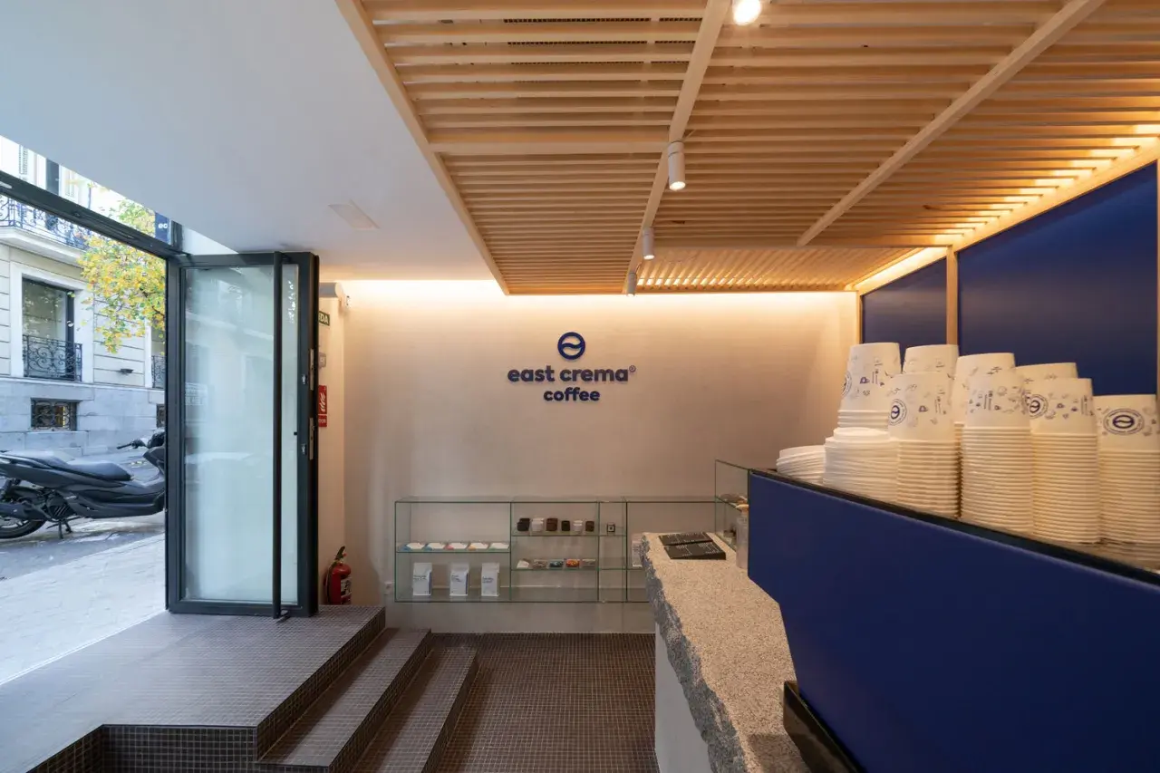 Interior de cafetería con logo "east crema coffee", vasos apilados y estanterías con productos.