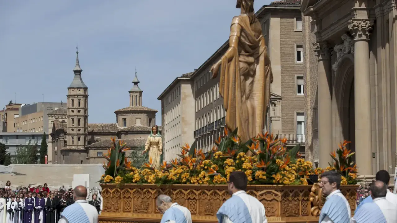 Procesión de **domingo de resurrección Zaragoza**. Figuras religiosas adornadas con flores en un paso, ante edificios históricos y una multitud.