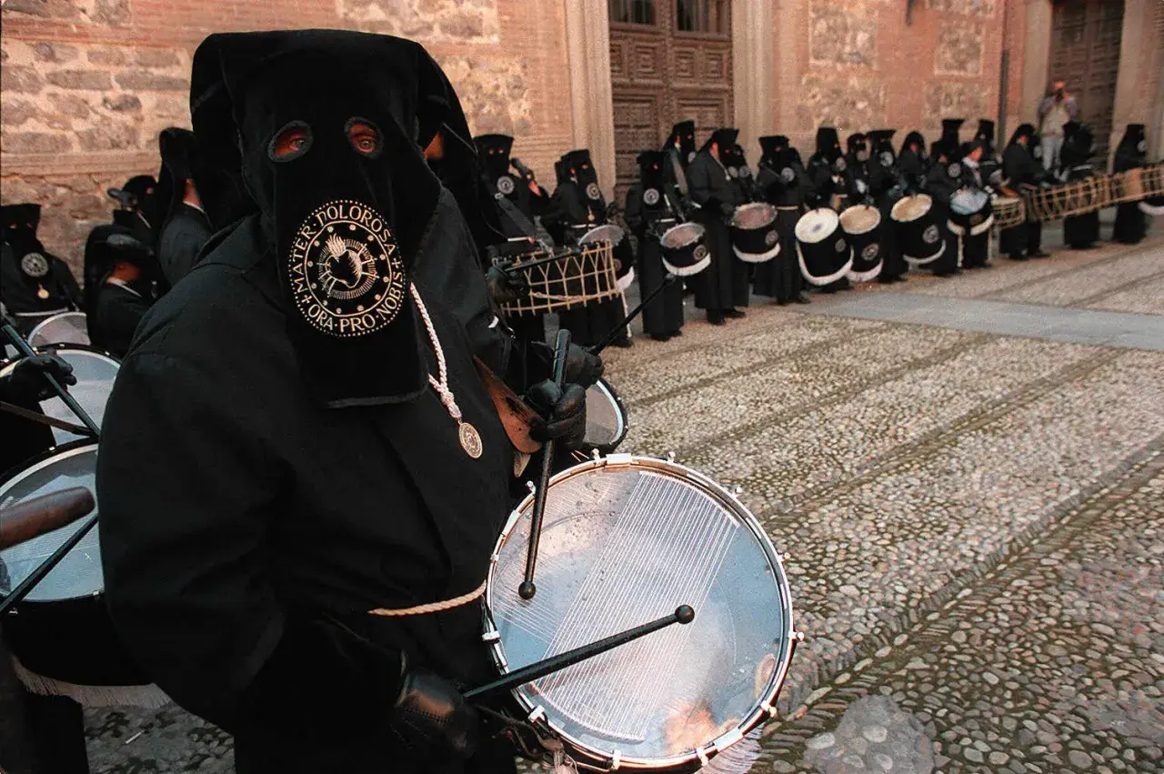 Tambores resuenan en las procesiones de Zaragoza. Nazarenos con túnicas negras y capirotes golpean sus tambores, creando un ritmo solemne.