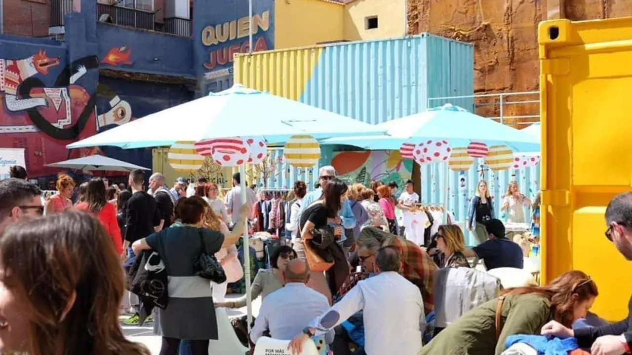Gente comprando ropa en un animado mercadillo de las armas, con coloridos contenedores y murales de fondo.
