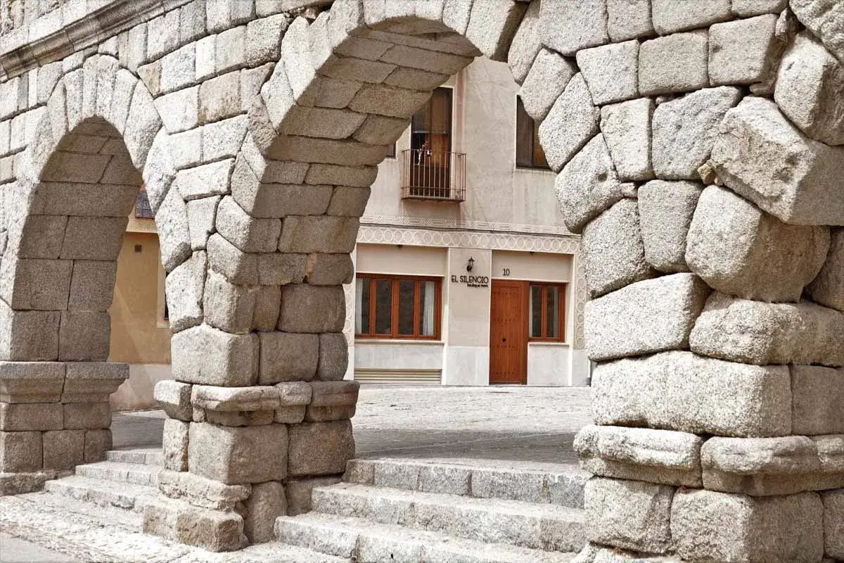 Arcos de piedra del Acueducto de Segovia, un lugar imprescindible que ver en Segovia. Al fondo, la fachada de un edificio con el letrero "El Silencio".