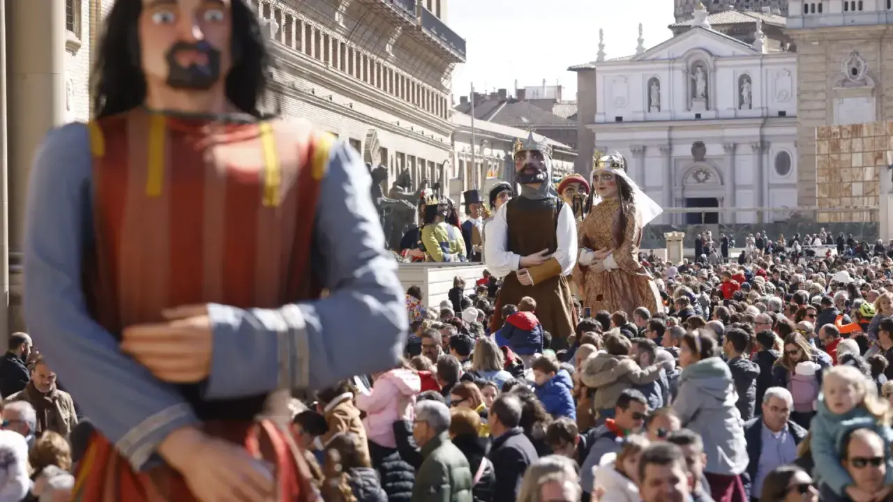 Gigantes y cabezudos desfilan entre una multitud en una plaza. Las figuras de cartón piedra, algunas con coronas, son el centro de atención.
