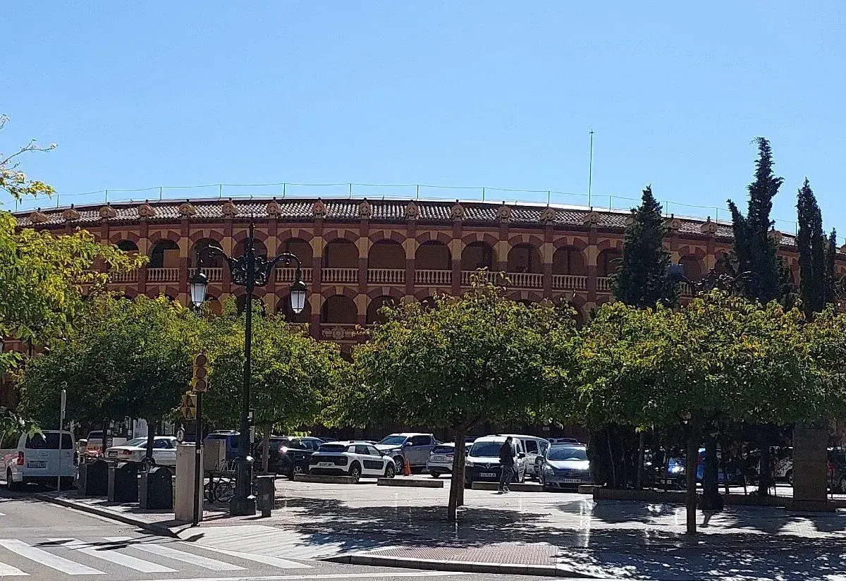 La plaza de toros de Zaragoza, con su arquitectura de arcos y balcones, se alza tras una hilera de árboles frondosos y un aparcamiento con varios coches.