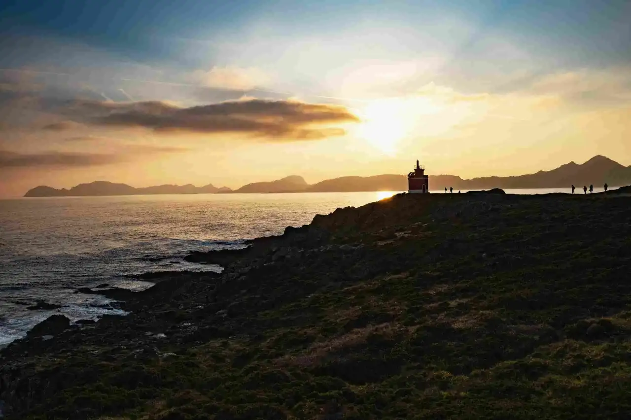 Atardecer en la costa gallega, con un faro solitario y siluetas de personas contemplando el mar. Un paisaje idílico para tus destinos Galicia.