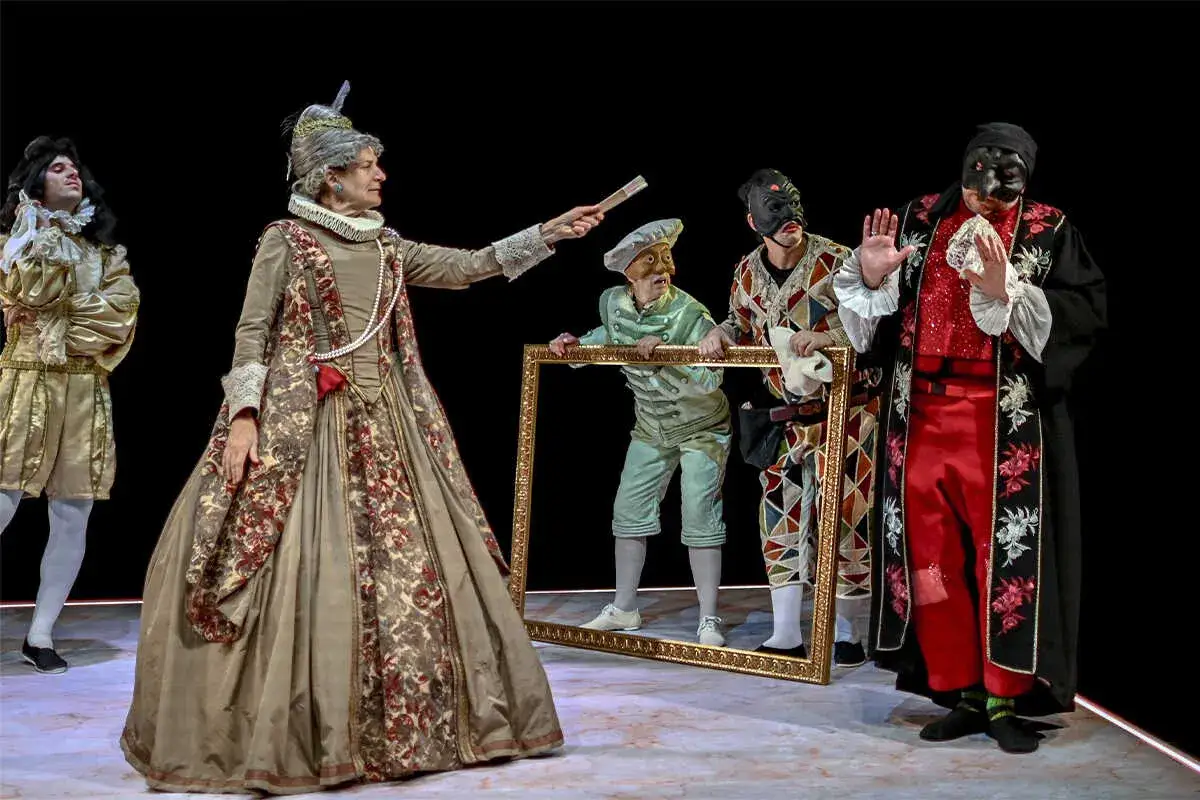 Escena teatral con personajes de els joglars zaragoza. Una mujer con vestido de época y abanico, un hombre con traje dorado, y otros con máscaras y ropajes coloridos.