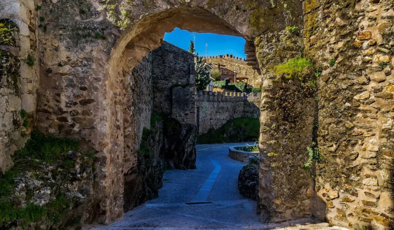 Antigua entrada de piedra con vegetación. Un camino empedrado serpentea hacia un pueblo fortificado, ideal para descubrir qué ver en la Comunidad de Madrid.