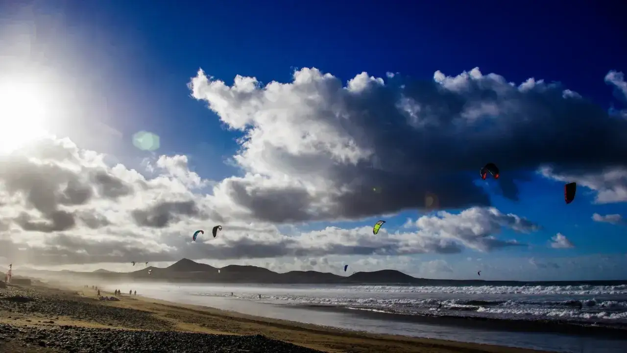 Deportistas disfrutan del kitesurf en Lanzarote bajo un cielo azul y nubes dramáticas.