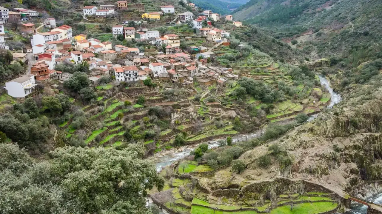 Pueblo en las Hurdes, con casas blancas y rojas escalonadas en la ladera, terrazas verdes y un río serpenteante.