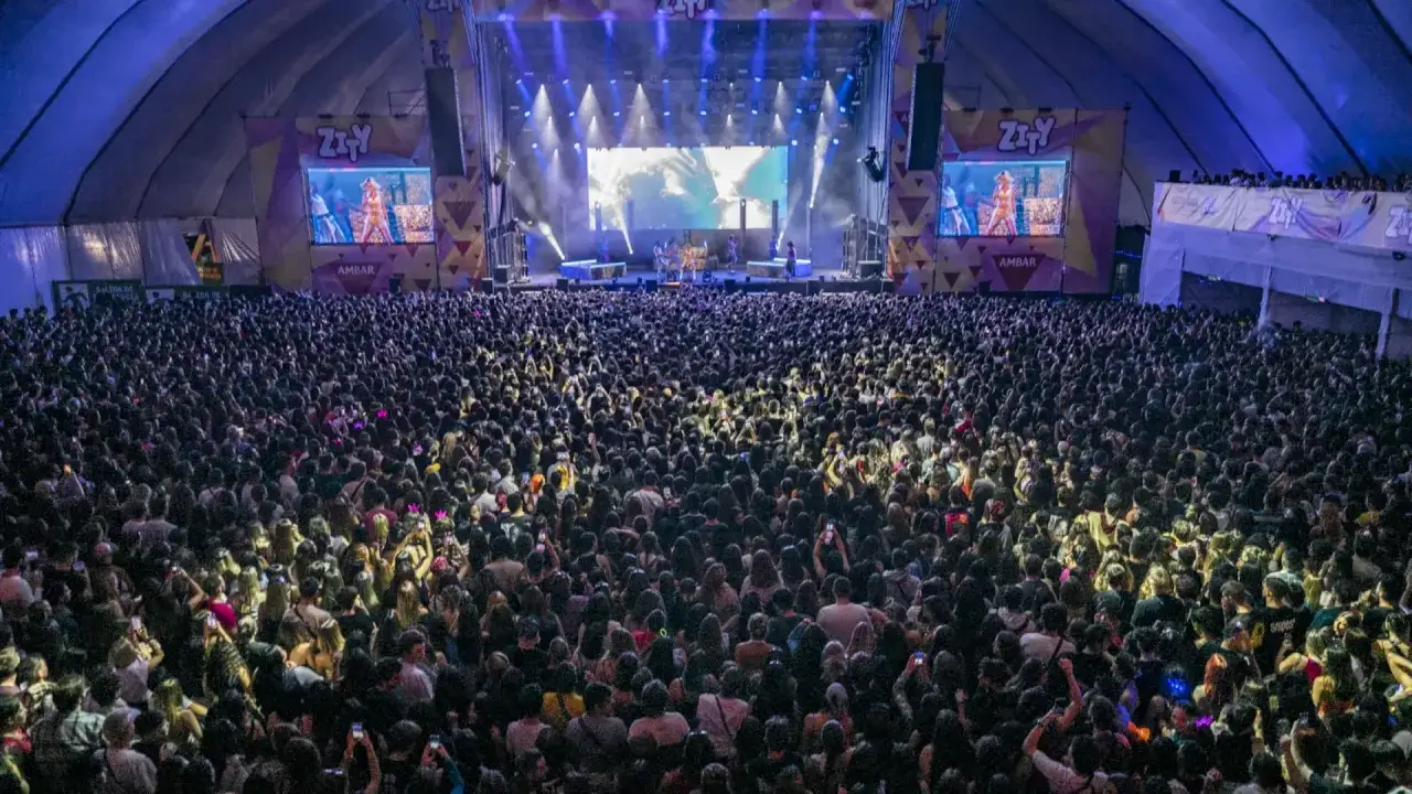 Multitud vibrante en un concierto bajo techo, con luces de escenario y pantallas gigantes mostrando el logo