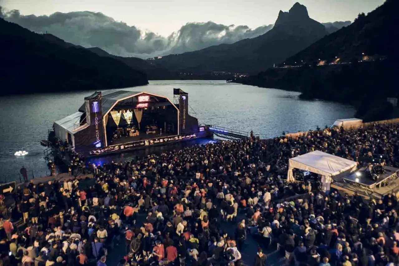 Gran multitud en un concierto al aire libre, con un escenario flotante en un lago y montañas al fondo. Noche de música en el concierto ZAZ Pirineos Sur.