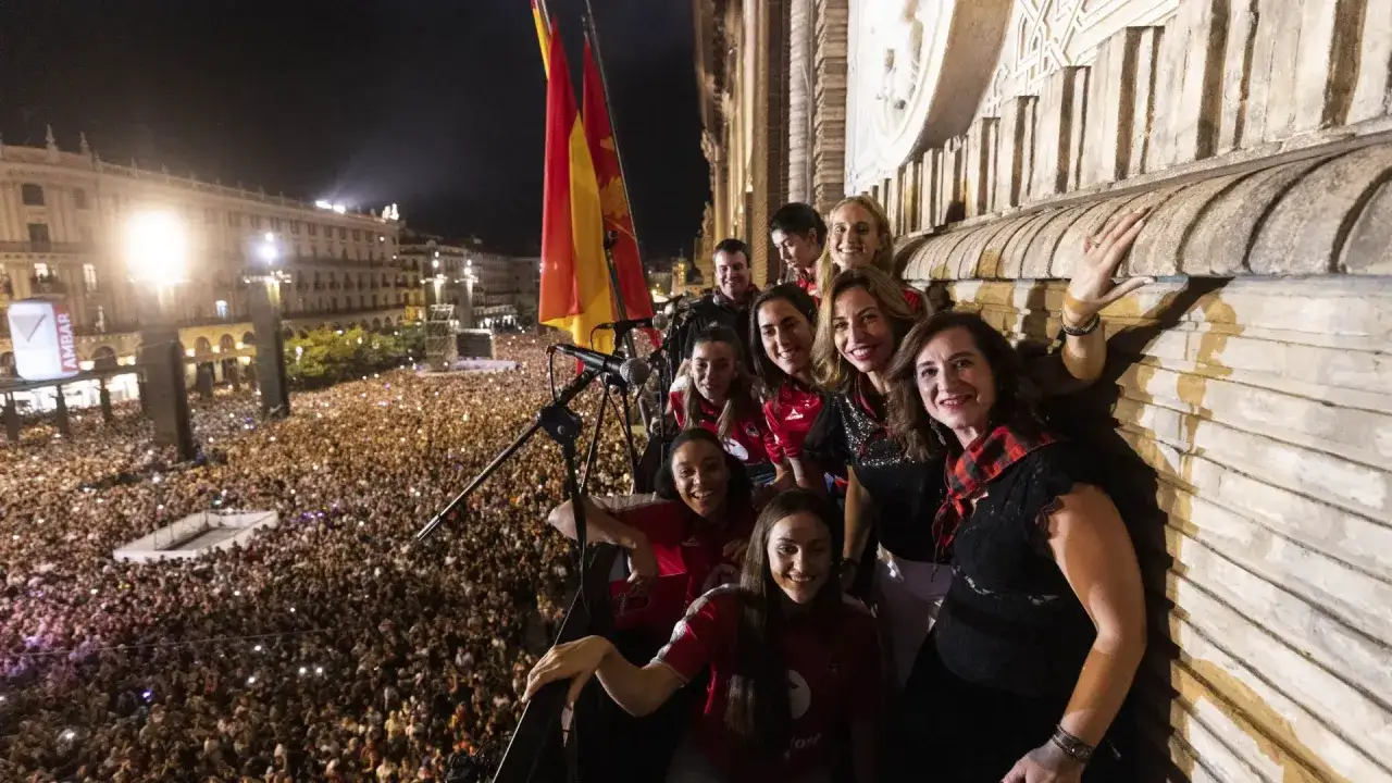 Un grupo celebra en un balcón con una multitud abajo. Las banderas ondean, anunciando el pregon de Pilares.