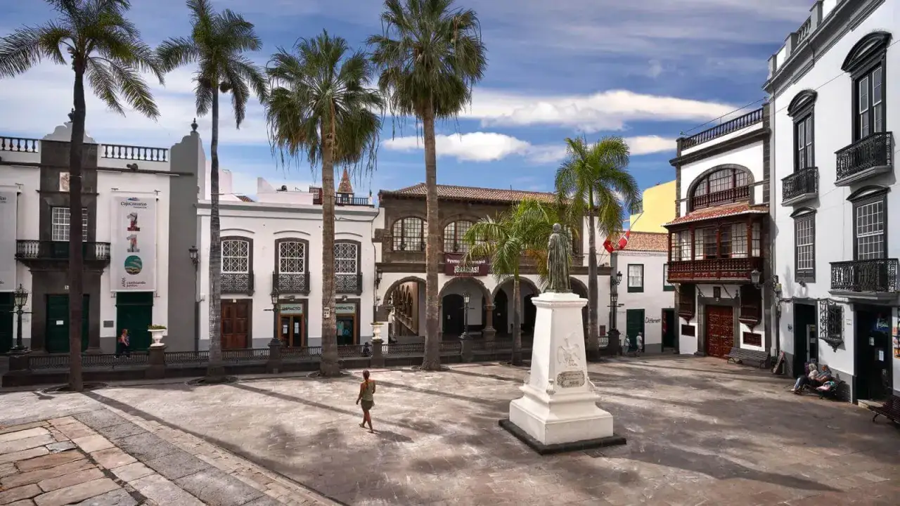 Plaza con palmeras y edificios coloniales, ideal para visitar La Palma. Una mujer pasea cerca de un monumento.