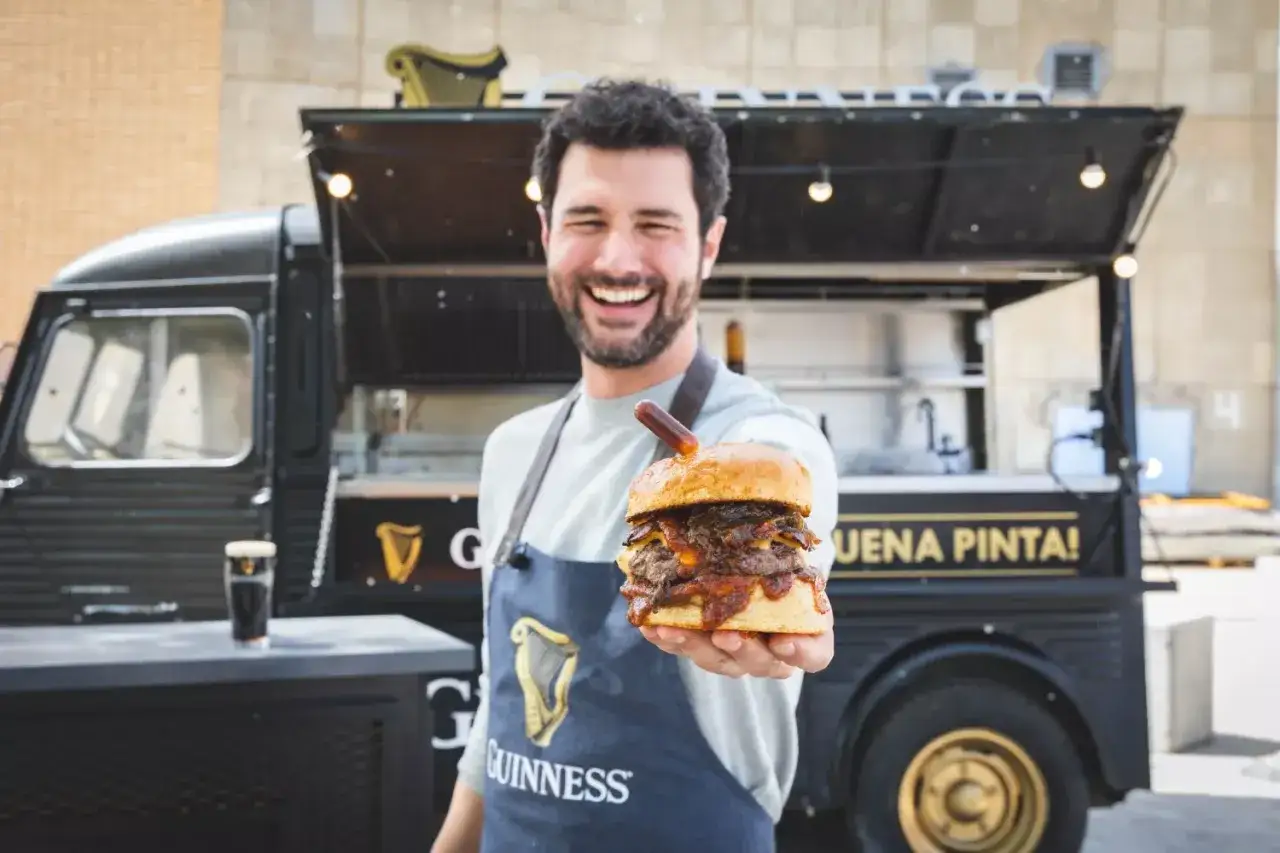 Hombre sonriente ofrece una jugosa hamburguesa en un food truck Guinness. ¡Un festín de sabor te espera!