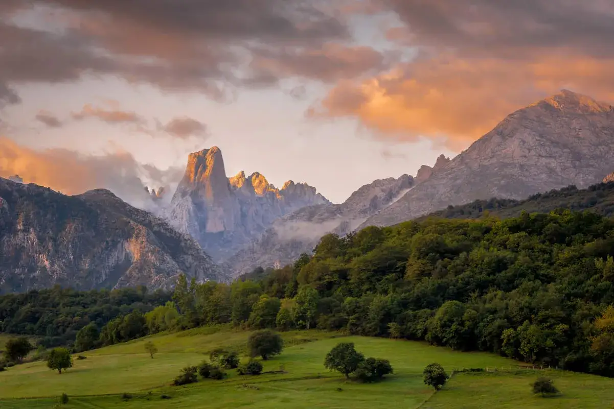 Majestuosas montañas iluminadas por el atardecer, un prado verde y árboles frondosos, un paisaje que evoca los lugares más bonitos de España.