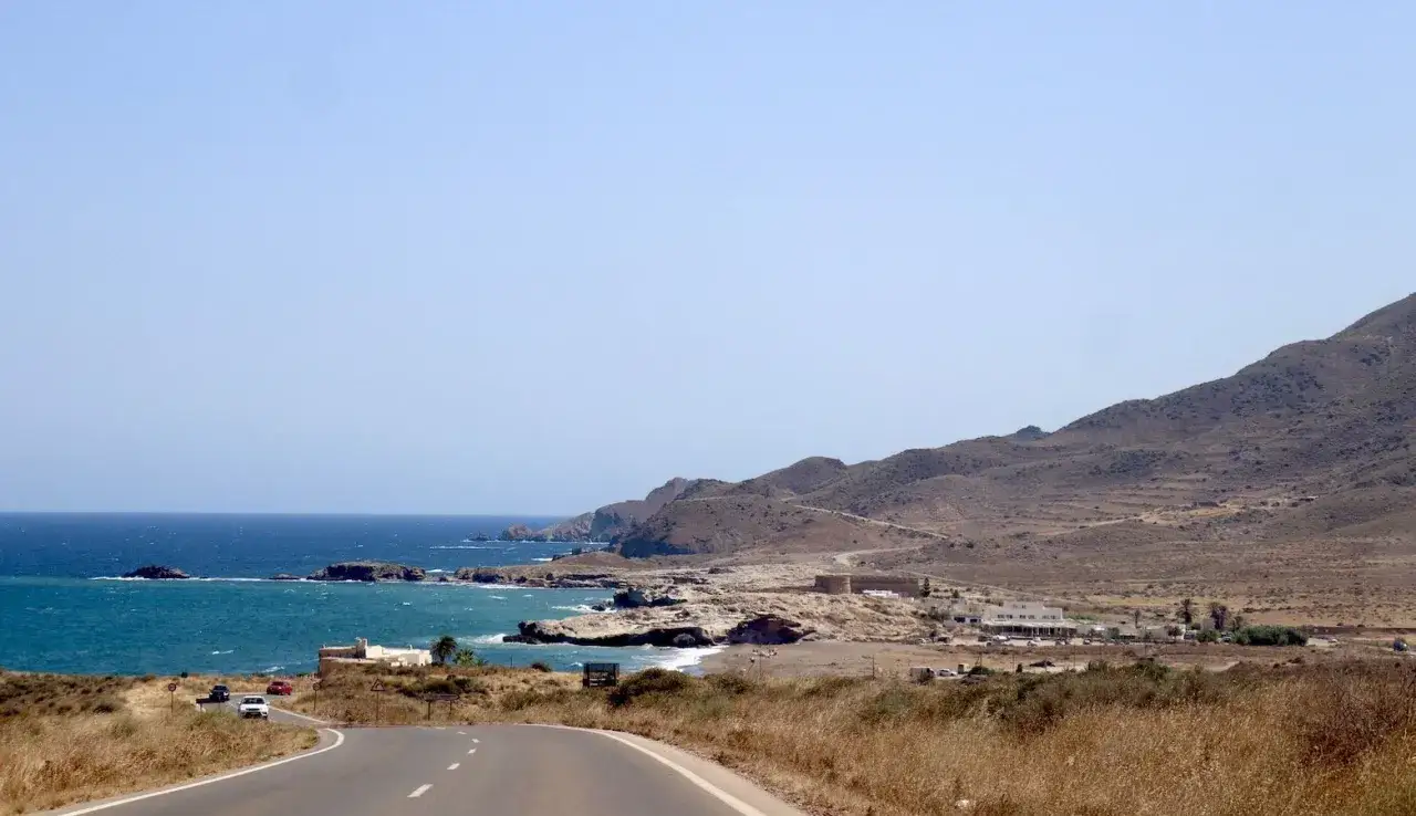 Carretera costera hacia Cabo de Gata, con mar azul, montañas áridas y edificios.