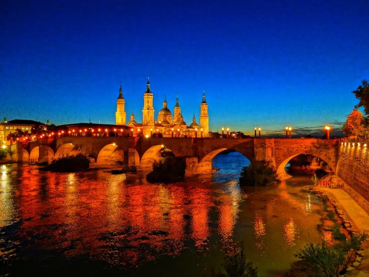 Un antiguo puente de piedra ilumina la noche con reflejos dorados sobre el río. Al fondo, una basílica majestuosa se alza bajo un cielo azul profundo.