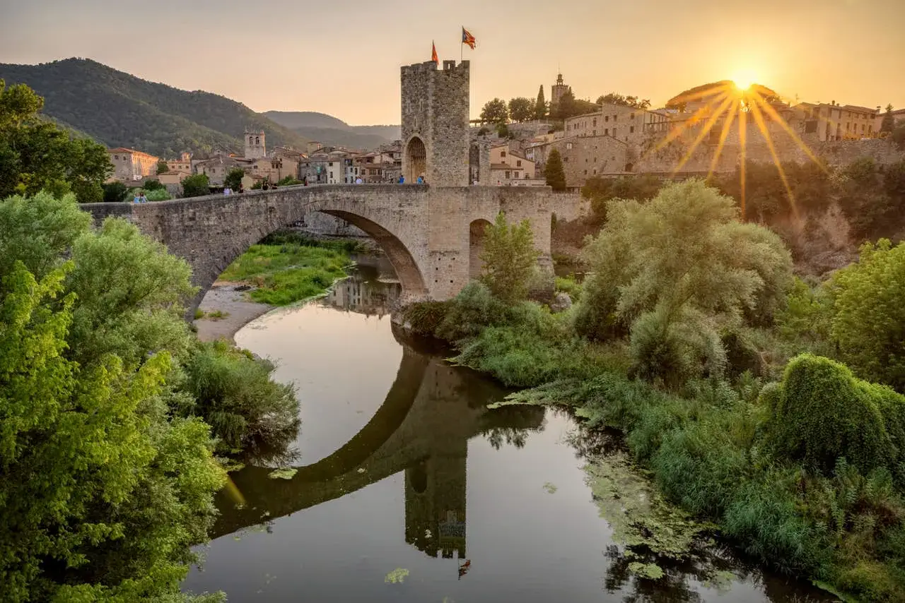 Un puente medieval con una torre fortificada, reflejado en un río sereno, bajo un atardecer dorado. Un ejemplo de los puentes de España que narran historias.