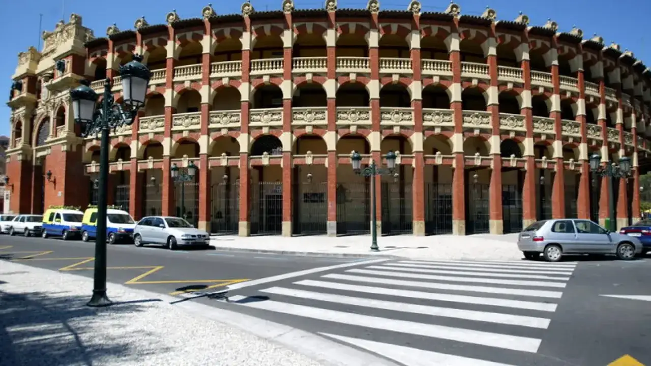 La plaza de toros de Zaragoza, con su arquitectura de ladrillo y arcos, se alza majestuosa. Coches aparcados y un paso de peatones completan la escena.