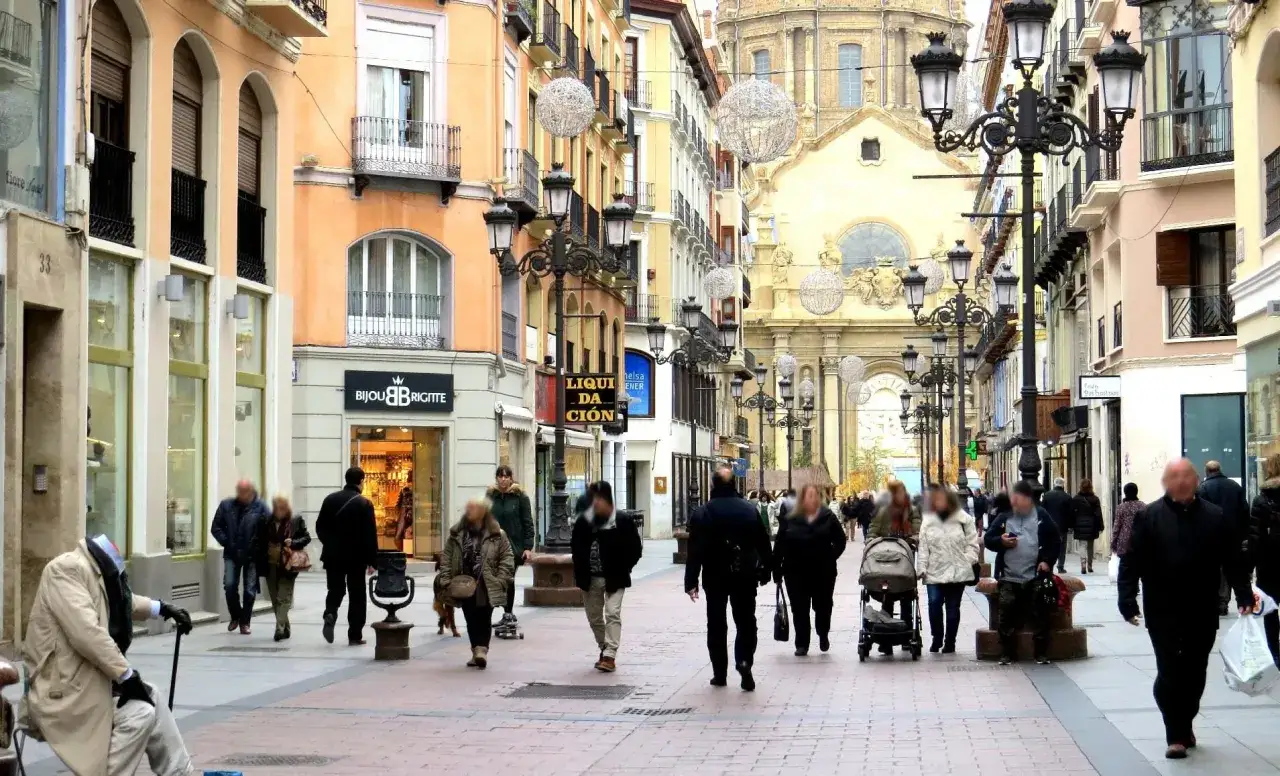 Gente pasea por una calle comercial con edificios hist&oacute;ricos y un gran templo al fondo, evocando el encanto de Zaragoza.