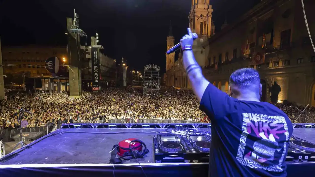 DJ animando una multitud masiva en las fiestas del Pilar, con conciertos y edificios históricos de fondo.