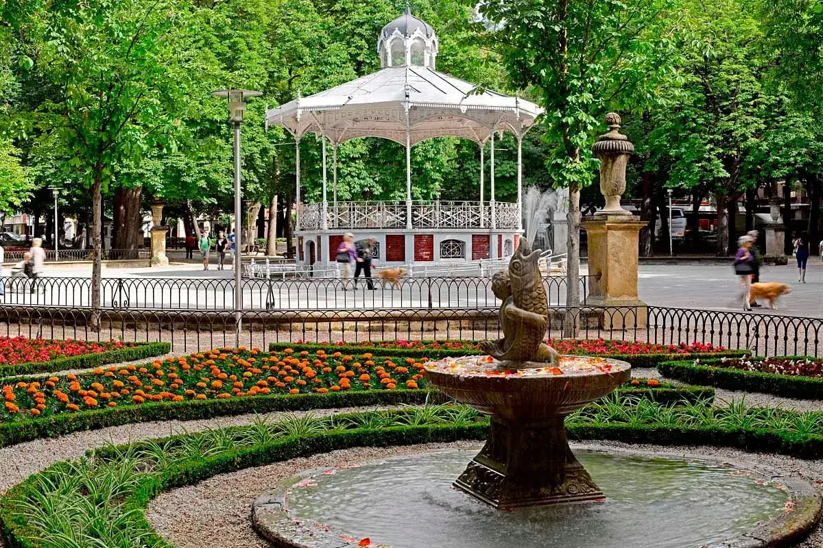 Fuente con pez y flores, ideal para ver en Vitoria. Kiosco blanco y gente paseando en un día soleado.
