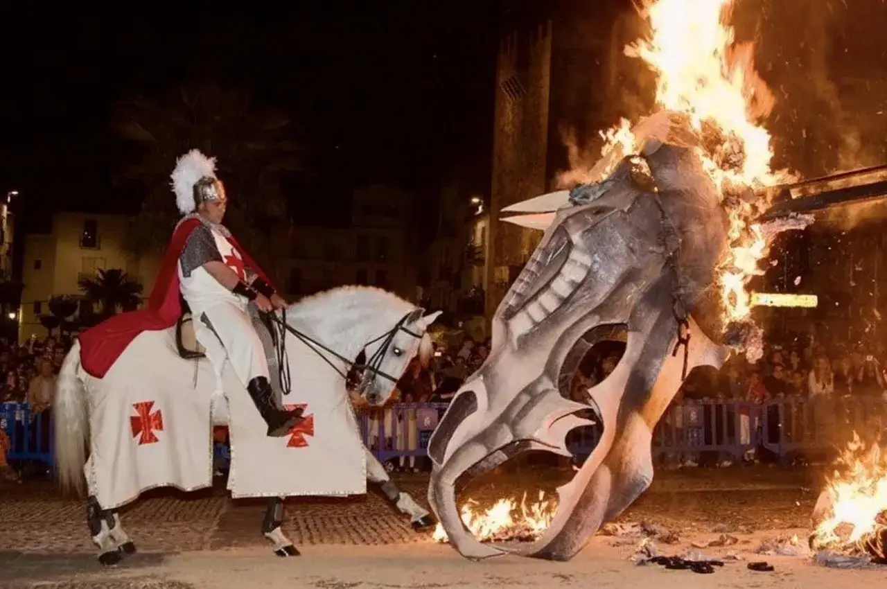 Caballero blanco, con capa roja y armadura, a caballo, se enfrenta a una cabeza de dragón en llamas. Representación de la historia de San Jorge.