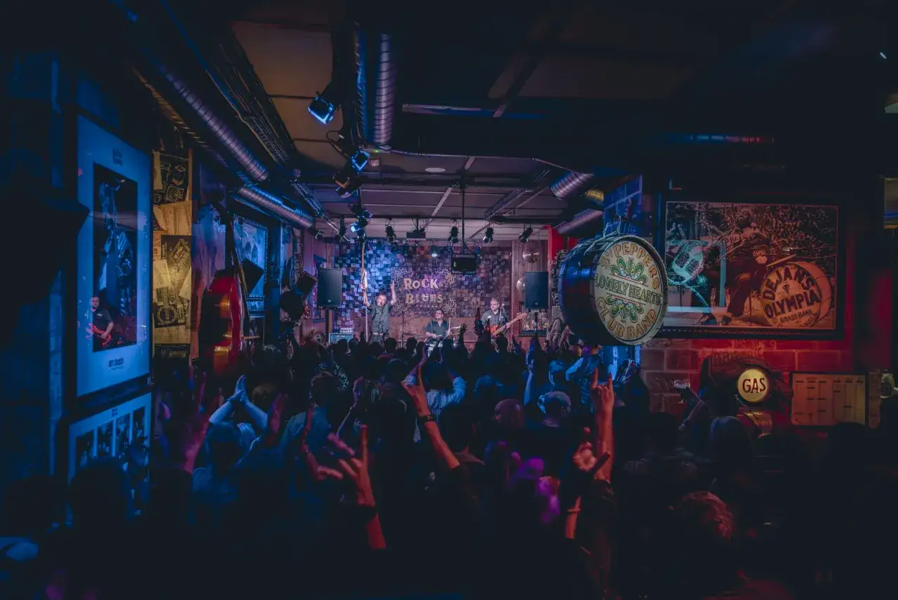 Una banda de rock y blues toca en un local con luces de neón. El público, con las manos en alto, parece disfrutar de la música, como si estuvieran en unas las vegas zaragoza de la música en vivo.