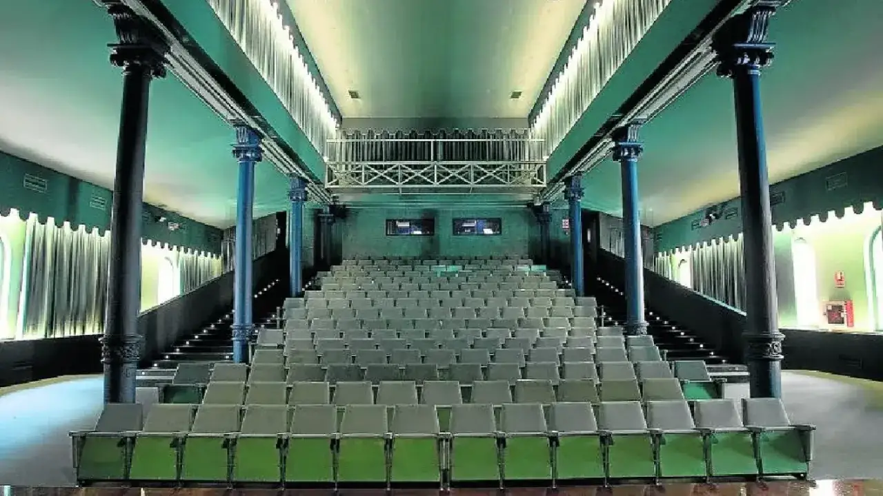 El interior del teatro Mercado de Zaragoza, con sus filas de asientos verdes y grises, columnas oscuras y una barandilla met&aacute;lica en el nivel superior.