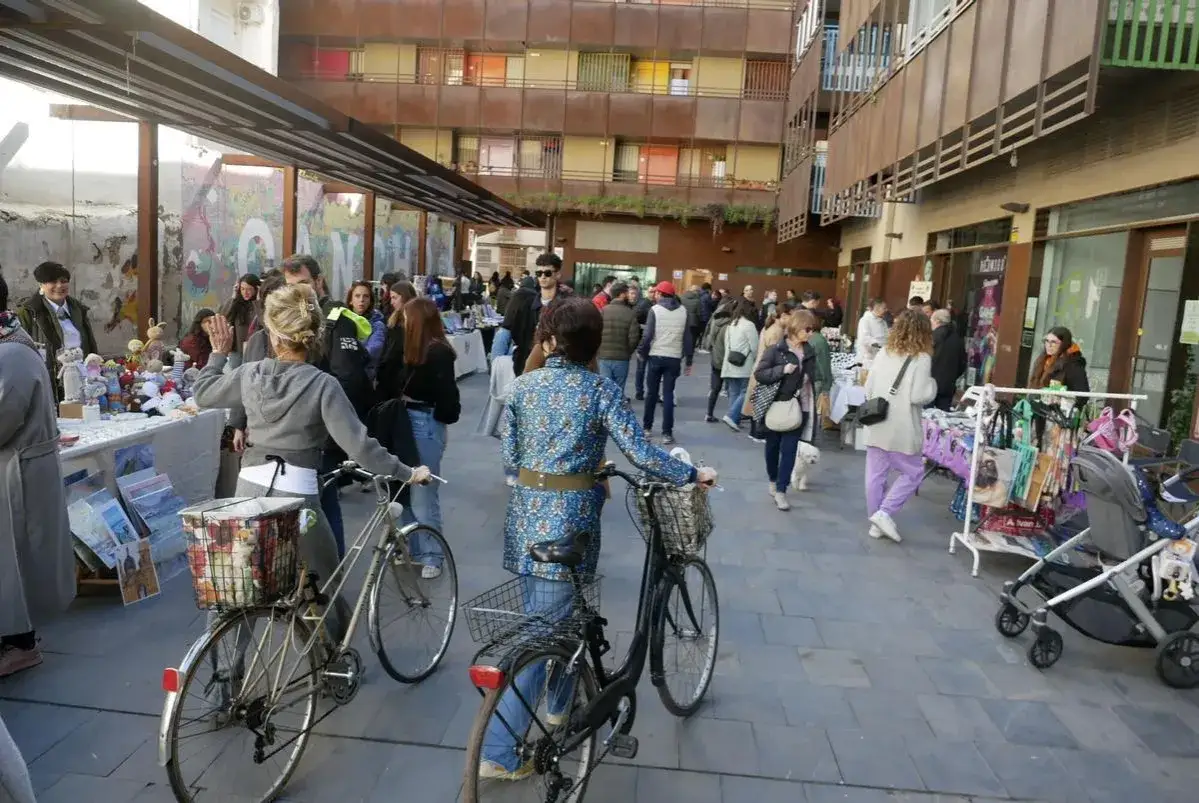 Gente pasea por un mercadillo de las armas, con puestos de artesanía y ropa. Hay bicicletas y un cochecito de bebé.