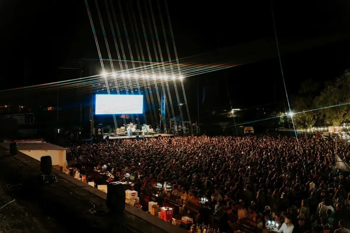 Multitud vibrante en un amante festival de música, con una banda tocando en el escenario iluminado y una gran pantalla de fondo.
