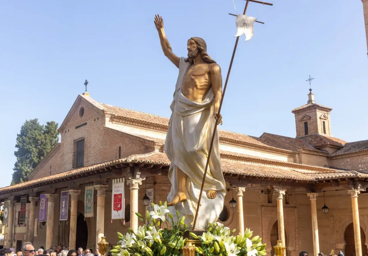 Imagen de una procesión del domingo de resurrección con una estatua dorada de Jesús resucitado, portando una bandera, adornada con lirios blancos, frente a una iglesia.