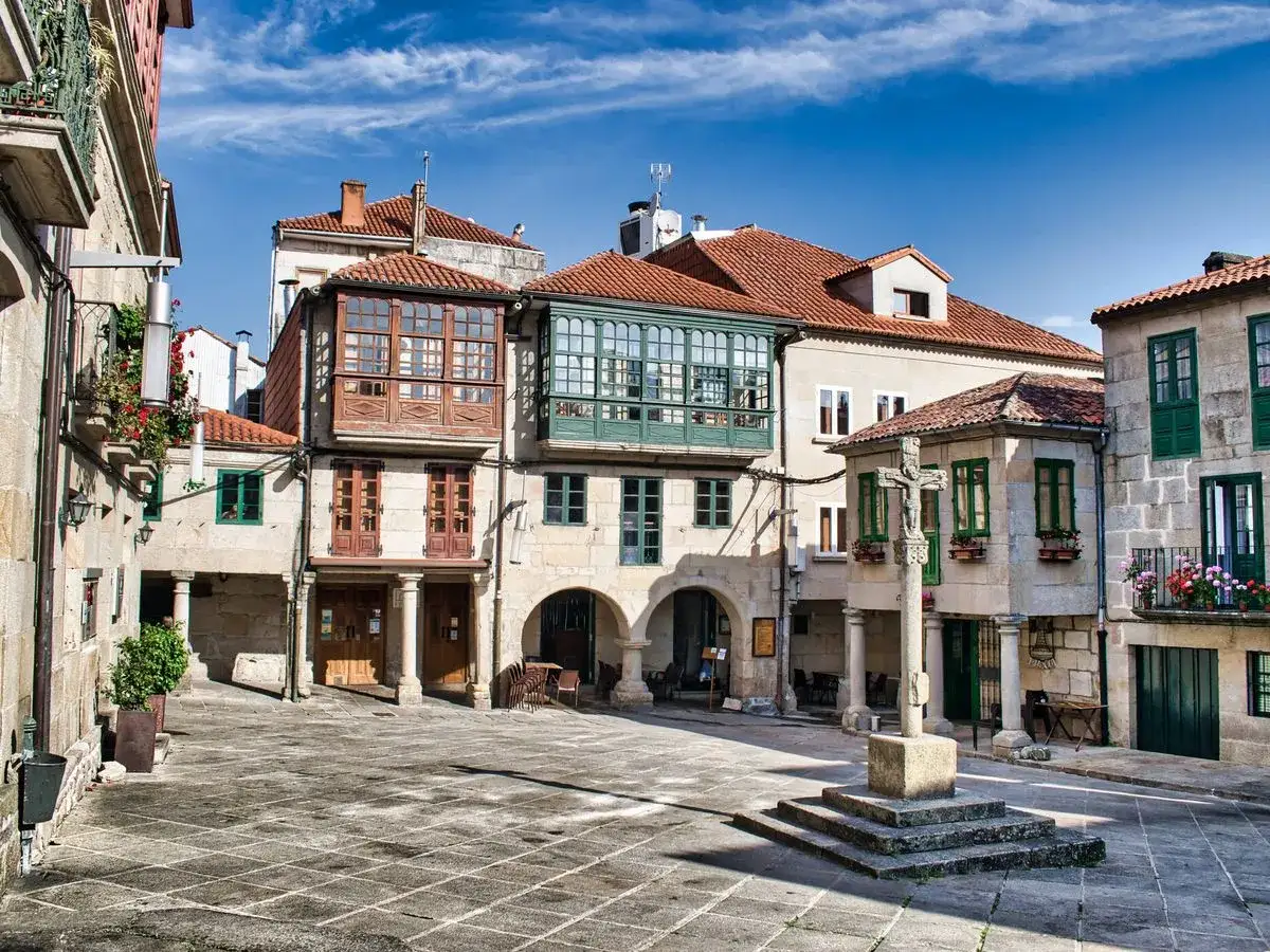 Plaza empedrada con cruceiro y edificios históricos, ideal para escapadas con encanto. Balcones de madera y ventanas verdes añaden carácter.