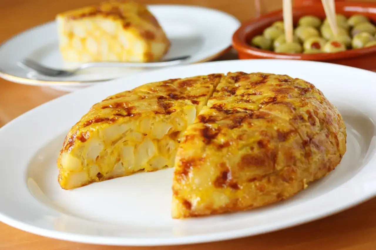 Tortilla de patata zaragoza, jugosa y dorada, servida en plato blanco. Al fondo, aceitunas y otra porción.