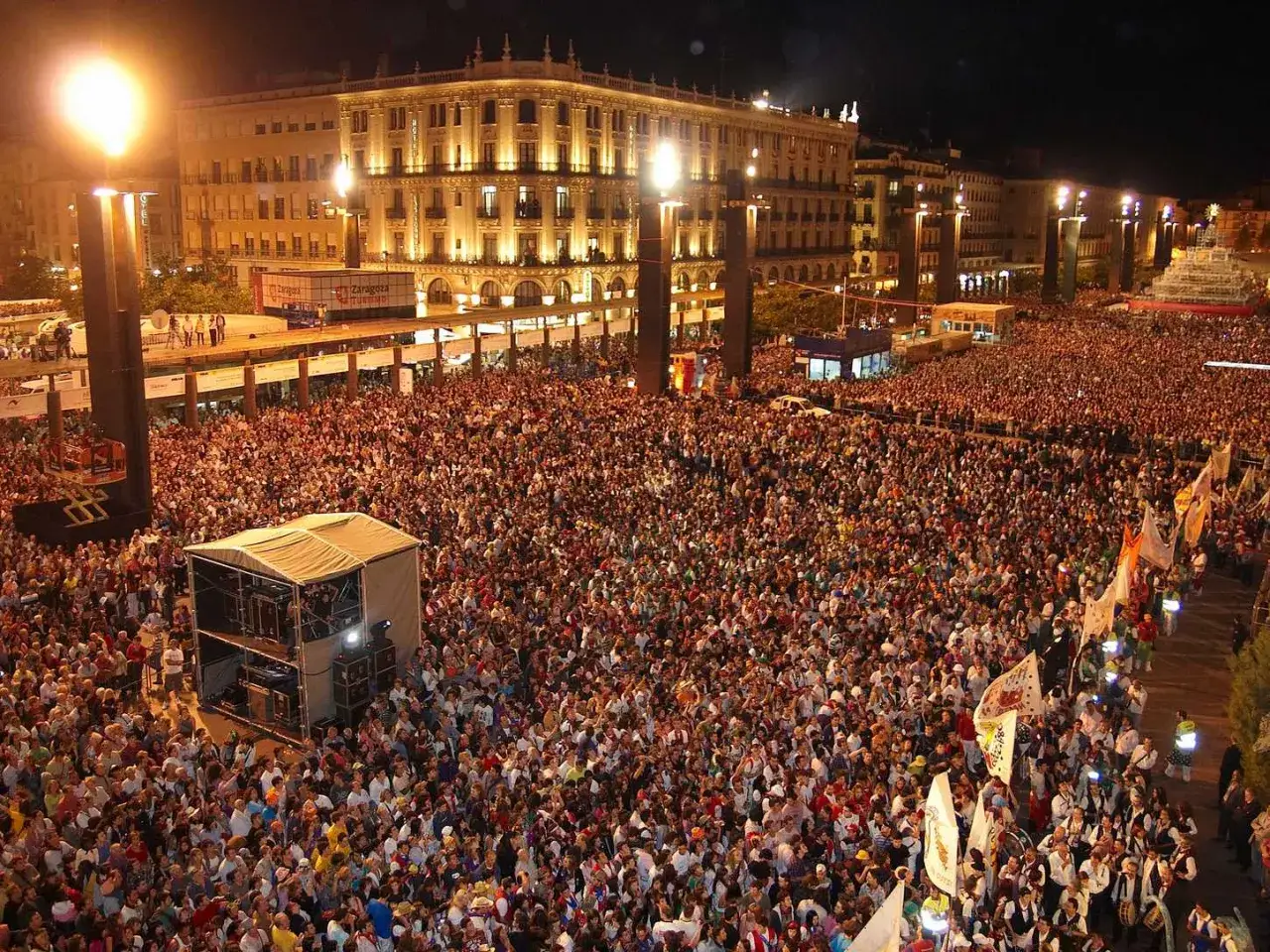 Multitudinaria celebración en las fiestas del barrio de Las Fuentes, Zaragoza. Gente reunida en una plaza iluminada por la noche, con un escenario y edificios históricos al fondo.