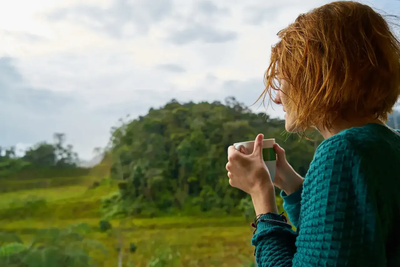 Mujer con cabello rojizo disfruta de una taza de té, contemplando el paisaje verde, ideal para un retiro espiritual en Cataluña.