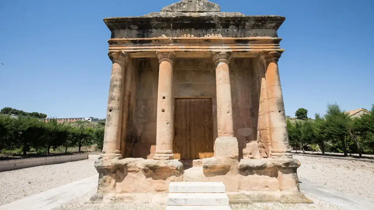 El mausoleo de Fabara, una estructura antigua con columnas y una puerta de madera, se alza bajo un cielo azul claro.